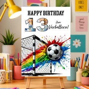 以下が含まれることがあります： 「HAPPY BIRTHDAY 13」の文字とサッカーをテーマにしたバースデーカード。数字の13はお金の写真で埋められています。サッカーボールは、カラフルなペイントスプラッシュデザインのゴールの前にあります。カードは小さな木製のイーゼルに表示されています。