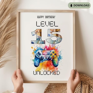 Op de afbeelding: Een ingelijste print met de woorden "Happy Birthday" en "Level 15 Unlocked". Het cijfer 15 is gevuld met een gelddesign. Onderaan staat een kleurrijke aquarel afbeelding van een videogamecontroller.