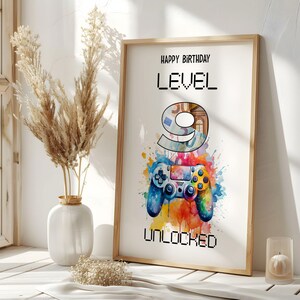 May include: A framed print with the text "HAPPY BIRTHDAY LEVEL 9 UNLOCKED". The artwork features a watercolour-style image of a video game controller with colourful paint splatters. A vase with dried plants is to the left.