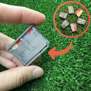 Puede incluir: Un soporte gris para tarjetas SD con pestañas de colores, sostenido en una mano. Varias tarjetas SD son visibles en un círculo en el fondo. El soporte está diseñado para organizar y almacenar tarjetas SD.