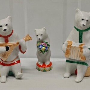 Puede incluir: Tres figuras de osos de cerámica blanca. El oso de la izquierda toca un instrumento de cuerda color canela y lleva adornos rojos. El oso central sostiene un ramo de flores coloridas sobre una base roja. El oso derecho toca un acordeón.