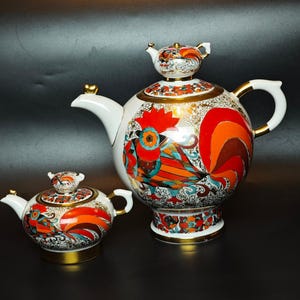 May include: A white porcelain teapot and a matching sugar bowl, both featuring a vibrant, multi-colored rooster design in shades of red, orange, and teal with gold accents. The teapot has a lid topped with a small rooster figure.