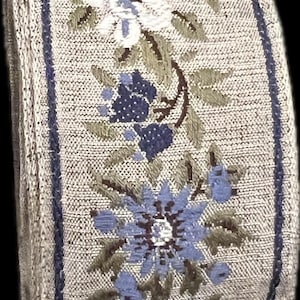 Può includere: Un rotolo di nastro decorativo con un motivo floreale. Il nastro presenta fiori ricamati in bianco e blu, con foglie verdi, su uno sfondo neutro. Bordi blu scuro incorniciano i bordi.