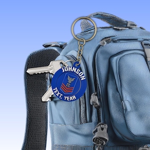 May include: A blue keychain with a gold ring and chain, featuring the name "JOHNSON" and the words "TEXT, YEAR". The keychain has a military insignia and is attached to a blue backpack with keys.