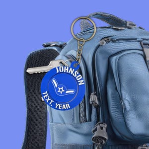 May include: A blue keychain with a gold ring and chain, featuring the name "JOHNSON" and the words "TEXT, YEAR" around a stylized emblem. The keychain is attached to a blue backpack with a key.