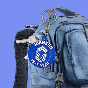 May include: A blue keychain with a gold ring and chain, featuring the name "JOHNSON" and the text "TEXT, YEAR". The keychain has a military insignia design. The keychain is attached to a blue backpack with keys.