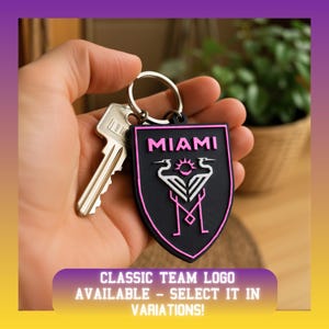 May include: A black shield-shaped keychain with a pink border featuring the text MIAMI in pink, a pink sun icon, and two white stylized herons facing each other above a pink M letter. A silver metal key ring is attached to a standard silver key.