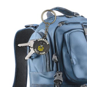 Puede incluir: Un llavero personalizado negro y dorado con el nombre "JOHNSON", texto y año. El llavero está sujeto a una mochila azul con llaves. El diseño es de inspiración militar con estrellas y rayas.