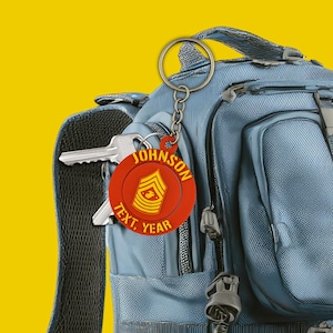 May include: A red and yellow keychain with the name "JOHNSON" and the text "TEXT. YEAR" printed on it. The keychain is attached to a silver keyring and is hanging on a blue backpack. The keychain has a military insignia.