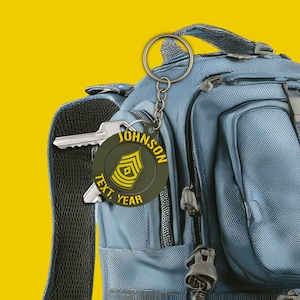 May include: A military-themed keychain with a circular tag featuring a yellow and green insignia and the name "JOHNSON". The keychain is attached to a blue backpack and includes a key. The tag also has the words "TEXT, YEAR".