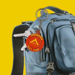 Puede incluir: Un llavero rojo y amarillo con el nombre "JOHNSON" y el texto "TEXT, YEAR" está sujeto a un llavero plateado. El llavero cuelga de una mochila azul. Las llaves son visibles en el bolsillo de la mochila.