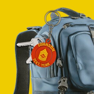 May include: A red and yellow keychain with the name "JOHNSON" and the text "TEXT, YEAR" is attached to a blue backpack. The keychain has a military insignia. A silver key is also attached to the backpack.