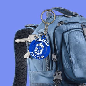May include: A blue keyring with a gold ring and chain, featuring the name "JOHNSON" and the text "TEXT, YEAR" around a military emblem. The keyring is attached to a backpack with keys.