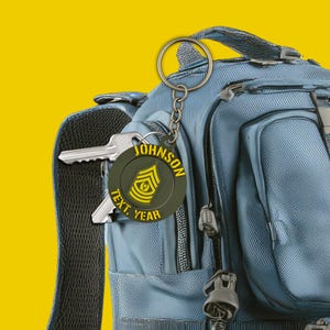 Puede incluir: Un llavero redondo verde oliva con texto amarillo y una insignia militar está sujeto a una mochila azul. El llavero dice "JOHNSON" en la parte superior, con "TEXT, YEAR" en la parte inferior. También hay una llave plateada.