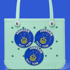 Può includere: Una borsa tote verde chiaro con tre toppe circolari blu e gialle. Ogni toppa ha il nome "JOHNSON", le parole "TEXT, YEAR" e una grafica. La borsa ha un manico e il marchio "bogg".