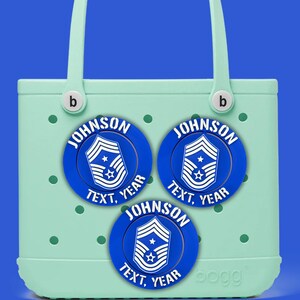 May include: A pale green tote bag with three blue circular patches. Each patch features the name "JOHNSON", the text "TEXT, YEAR", and a military insignia. The bag has a handle and the brand name "bogg".