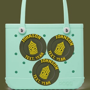Puede incluir: Un bolso tote verde claro con tres parches circulares. Cada parche tiene una insignia militar amarilla y verde oliva con el texto "JOHNSON" arriba y "TEXT, YEAR" abajo. El bolso tiene un asa y la marca "bogg" es visible.