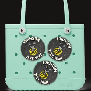 Pode incluir: Uma sacola verde-claro com três patches circulares pretos e amarelos. Cada patch tem o nome "JOHNSON", o texto "TEXT, YEAR" e um emblema de estilo militar. A sacola tem alças brancas e a marca "bogg".