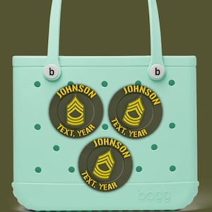 Puede incluir: Un bolso tote de color verde azulado claro con tres parches circulares. Cada parche tiene una insignia militar amarilla y negra, el nombre "JOHNSON" y las palabras "TEXT, YEAR". El bolso tiene asas blancas y la marca "bogg" es visible.