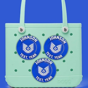 Peut inclure: Un sac fourre-tout vert clair avec trois écussons circulaires bleus. Chaque écusson porte le nom "JOHNSON", le texte "TEXT, YEAR" et un emblème blanc. Le sac a des poignées blanches et la marque "bogg".