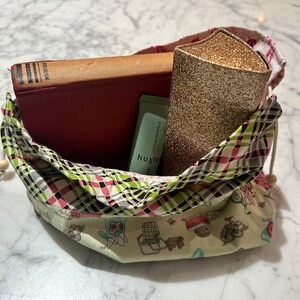 May include: A fabric drawstring pouch featuring a cream base with a pattern of small chairs and a plaid rim in green, pink, and black. Inside the pouch are a red hardcover book, a light green tube labelled Lemon Myrtle, and a gold glittery case.