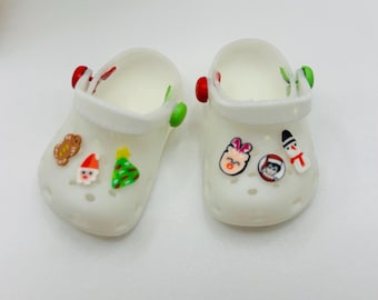 May include: Two white miniature clogs with Christmas-themed charms. One clog features a gingerbread man, Santa, and a Christmas tree. The other clog has a pink bunny, a character in a hat, and a snowman. The clogs have red and green accents.