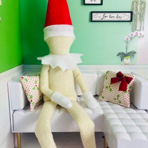 May include: A white elf doll wearing a red and white Santa hat sits on a white couch with a red and green patterned pillow. The couch is in a room with green walls and signs that say "Welcome Home", "Stay Awhile", and "Love Lives Here".