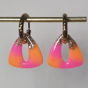 Peut inclure: Une paire de boucles d'oreilles créoles en métal doré texturé avec des pendentifs triangulaires en résine. Les pendentifs présentent un dégradé vif de rose fuchsia et d'orange, agrémenté d'une touche de paillettes argentées au sommet.