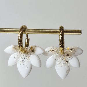 Pode incluir: Um par de brincos de argola dourados com textura martelada, cada um com um pingente branco em forma de pétala. Os pingentes são decorados com glitter dourado na base, criando um design inspirado em uma flor.