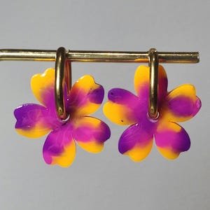 Peut inclure: Une paire de boucles d'oreilles en forme de fleur aux couleurs vives. Les pétales sont un dégradé de jaune et de violet, fixés à des créoles dorées. Les boucles d'oreilles sont présentées sur un fond gris neutre.