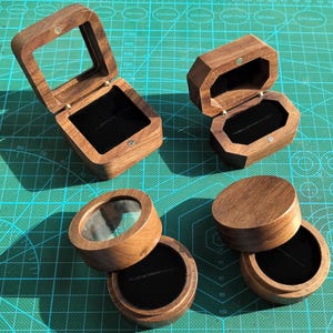 Puede incluir: Una colección de joyeros de madera de varias formas. Las cajas son de madera marrón claro y tienen un interior de terciopelo negro. Algunas cajas tienen tapas transparentes, otras tapas de madera maciza.