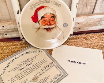 May include: A white ceramic plate with a festive Christmas design featuring Santa Claus. The plate has the text "Christmas 1988" and a certificate of authenticity with the text "Santa Claus".