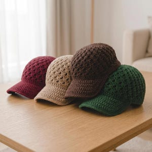Può includere: Quattro cappellini da baseball all'uncinetto in fila, ognuno di un colore diverso. I colori sono bordeaux, beige, marrone e verde. I cappellini sono realizzati in un materiale strutturato a trama aperta e poggiano su una superficie di legno marrone chiaro.
