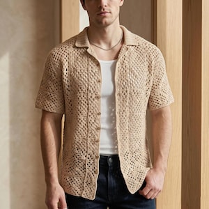 May include: A beige crochet shirt with short sleeves and a collared neckline. The shirt has a button-down front and a pocket on the left chest. The shirt is worn over a white tank top and paired with dark blue jeans. The shirt's open weave design is visible.