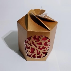 Puede incluir: Una caja de regalo hexagonal dorada con un recorte decorativo que revela un interior rojo. La caja tiene una parte superior plegada única y está diseñada para pequeños regalos o recuerdos. El recorte presenta un intrincado diseño floral.