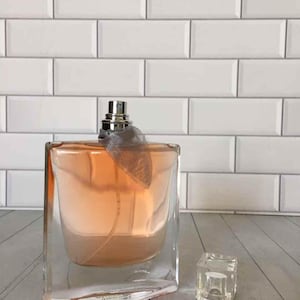 May include: A clear glass perfume bottle with a peach-colored liquid inside. The bottle has a silver sprayer and a decorative gray ribbon. A small, clear, square cap sits beside the bottle. The background is a white brick wall.