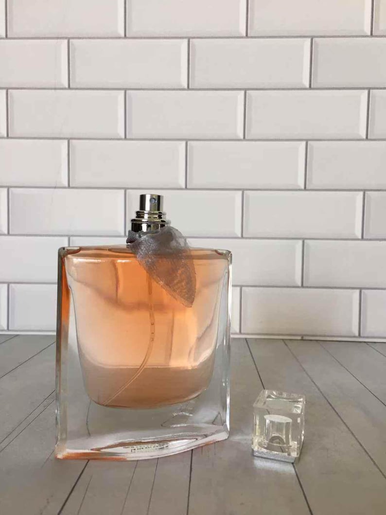 May include: A clear glass perfume bottle with a peach-colored liquid inside. The bottle has a silver sprayer and a decorative gray ribbon. A small, clear, square cap sits beside the bottle. The background is a white brick wall.