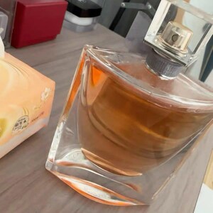 May include: A clear glass perfume bottle with a square, transparent cap. The bottle contains a light brown liquid. The perfume bottle is angled on a wooden surface, with a box of tissues and other items in the background.