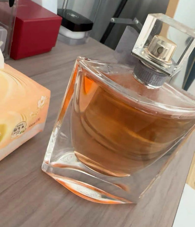 May include: A clear glass perfume bottle with a square, transparent cap. The bottle contains a light brown liquid. The perfume bottle is angled on a wooden surface, with a box of tissues and other items in the background.