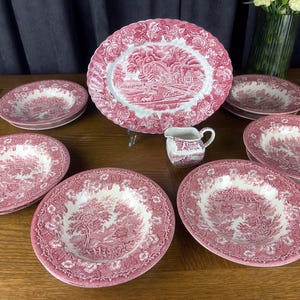 May include: A collection of pink and white ceramic dinnerware featuring a detailed pastoral transferware pattern. The set includes a large oval platter, several deep bowls, and a small creamer pitcher, all decorated with intricate floral borders.