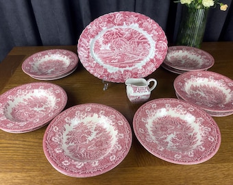 May include: A collection of pink and white ceramic dinnerware featuring a detailed pastoral transferware pattern. The set includes a large oval platter, several deep bowls, and a small creamer pitcher, all decorated with intricate floral borders.