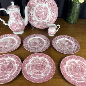 Puede incluir: Un juego de té de cerámica blanca con un patrón de transferencia rosa de paisajes rurales y bordes florales. El conjunto incluye una tetera con tapa, un plato grande para servir, una jarra pequeña para crema y seis platos a juego con escenas.