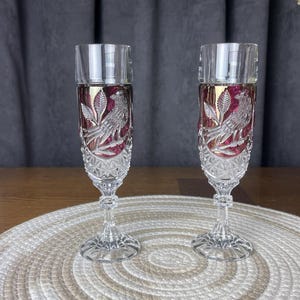 Puede incluir: Dos copas de champán de cristal transparente con un inserto de vidrio rojo intenso. Cada copa muestra un diseño grabado de un pájaro posado en una rama, un tallo facetado y un patrón de diamante texturizado en la base y parte inferior.