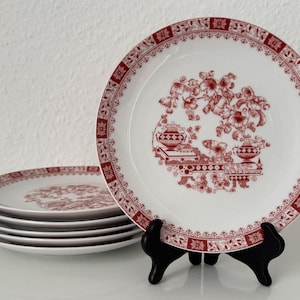 Può includere: Un set di piatti in ceramica bianca con un design floreale rosso. I piatti hanno un bordo decorativo e un motivo centrale di fiori e vasi. Un piatto è esposto su un supporto nero, mentre gli altri sono impilati. I piatti sono adatti per servire cibo.