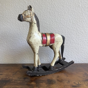 May include: A decorative wooden rocking horse featuring a distressed off-white finish with black accents on the mane, tail, and hooves. The horse includes a red and gold saddle detail and is mounted on a black curved base with a weathered appearance.