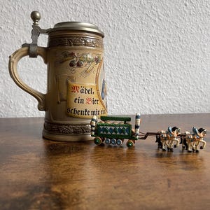 Może przedstawiać: Beżowy ceramiczny kufel do piwa z metalową pokrywką i uchwytem, ozdobiony reliefem przedstawiającym chmiel, pszenicę oraz zwój z niemieckim napisem "Mädel, ein Bier schenke mir ein". Obok znajduje się miniaturowy zielony wóz konny z dwoma końmi.