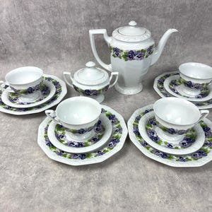 Op de afbeelding: Een wit porseleinen theeservies met een paars viooltjespatroon. De set bevat een theepot, een suikerpot met deksel en drie serviesdelen, elk bestaande uit een theekopje, een schoteltje en een groter bord met een geschulpte rand.