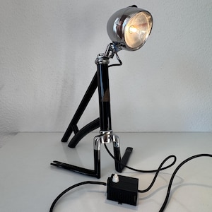 Op de afbeelding: Een tafellamp gemaakt van een zwart fietsframe en een verchroomde fietslamp. De lamp is voorzien van een zwart netsnoer met een rechthoekige zwarte schakelaar. De verchroomde koplamp bevindt zich aan de bovenzijde en geeft warm licht.