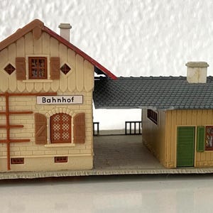 May include: A miniature model of a train station with the word "Bahnhof" on the front. The station has a beige facade with brown trim, a red roof, and a green side building. The model is set on a light-colored base.