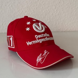Könnte beinhalten: Eine leuchtend rote Baseballkappe mit weißem Sticktext Deutsche Vermögensberatung, einem weißen kreisförmigen Logo mit Globus, fünf weißen Sternen, einer weißen Unterschrift auf dem Schirm und einem weißen quadratischen Aufnäher mit der Zahl 1 an der Seite.